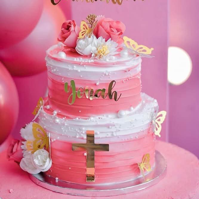 Two-tier pink and white gradient cake with a gold cross, pink roses, and gold butterflies