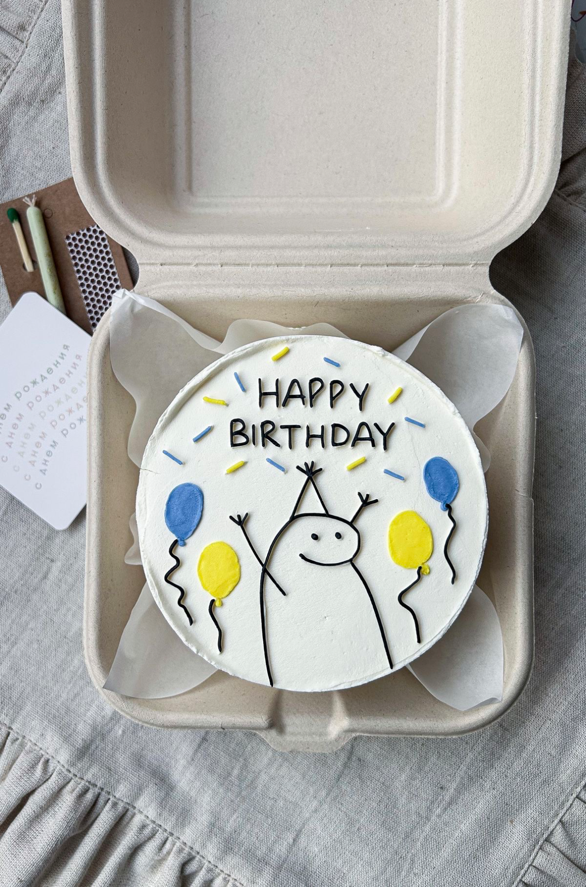 White bento cake with a hand-drawn Flork character wearing a party hat and holding yellow and blue balloons with Happy Birthday text.