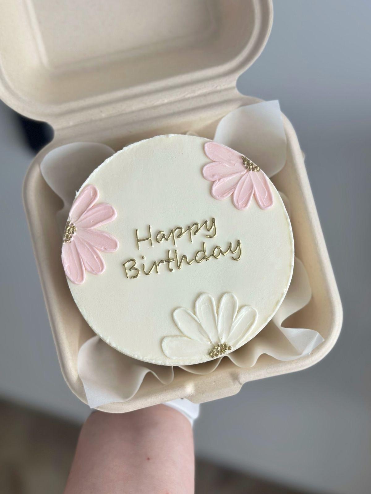 White bento cake with pink and white palette knife icing flowers and gold Happy Birthday message in a takeout box.