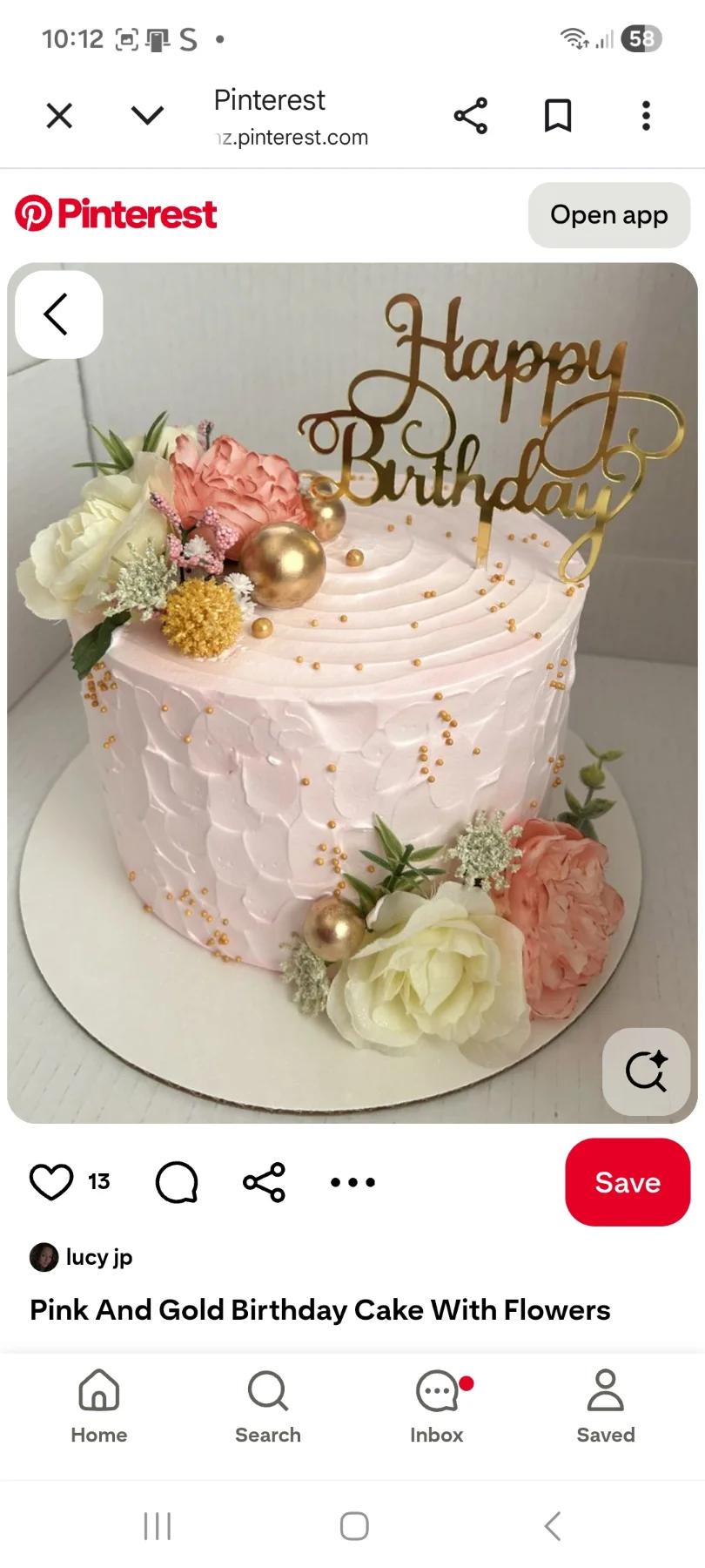 A single-tier light pink birthday cake with textured icing, gold dragees, artificial flowers, and a gold Happy Birthday topper.