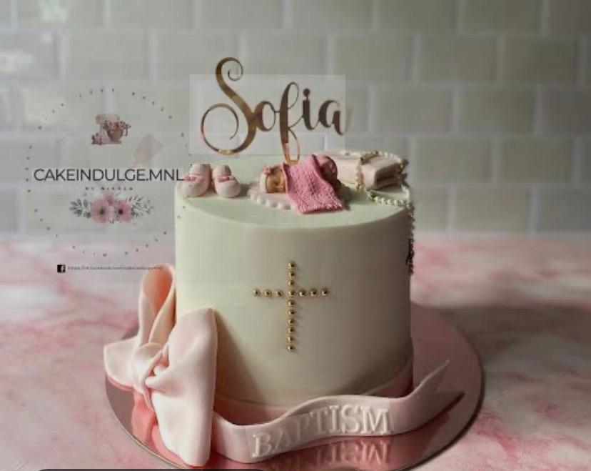 White baptism cake with a pink fondant bow, a gold cross made of beads, and a sleeping baby topper with pink accessories.